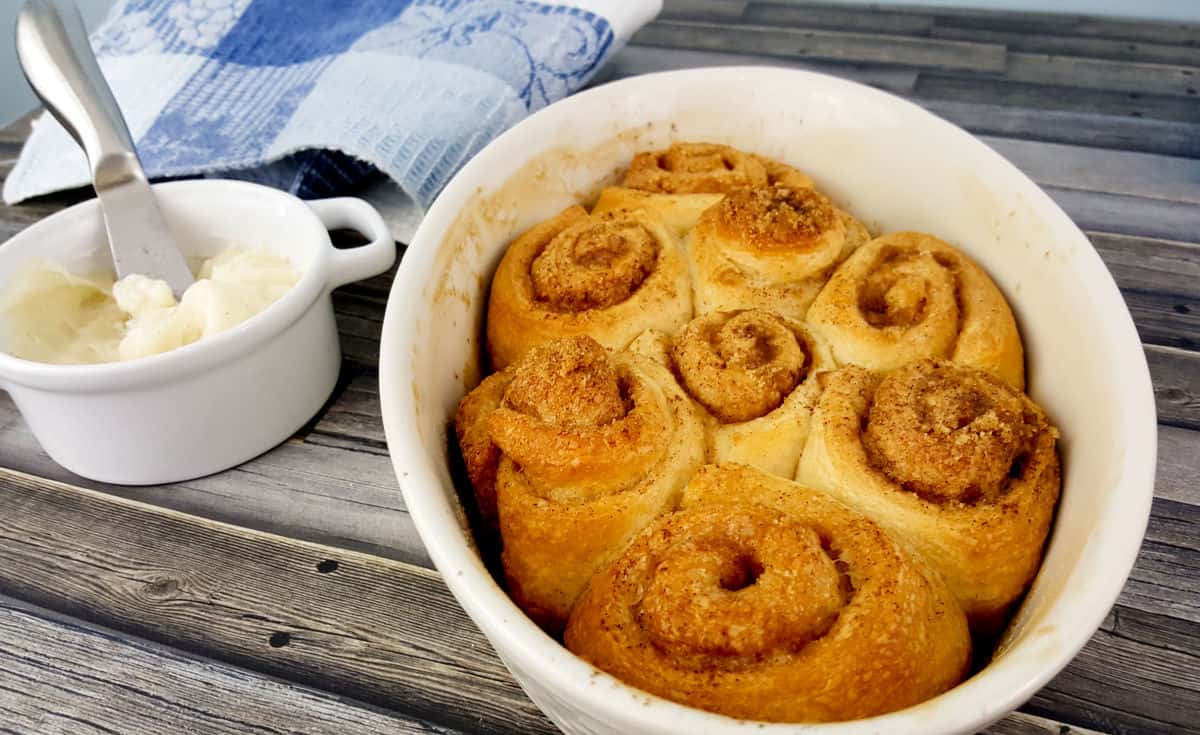 baked cinnamon rolls in a white dish next to a container of frosting. 