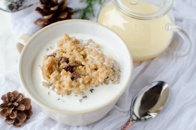 Crock Pot Eggnog Oatmeal in a white bowl next to a spoon, pinecones, and a pitcher of eggnog