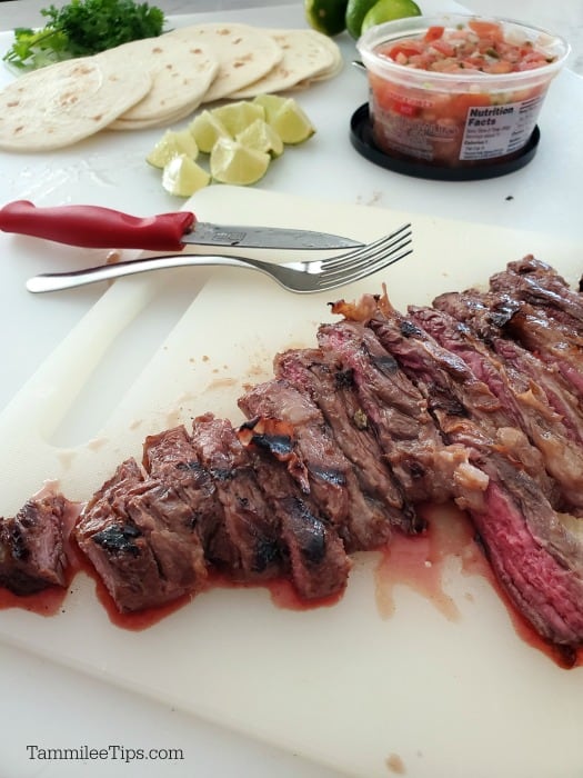 Carne asada cut into pieces on a cutting board next to salsa, lime wedges and soft street taco shells