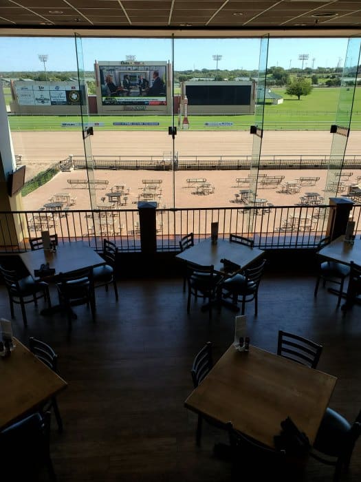 looking out at a race track with dirt track, tables and chairs and a large tv screen. 