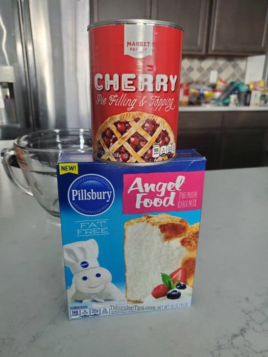 Can of cherry pie filling on top of  a box of angel food cake on a white kitchen counter with a bowl behind them