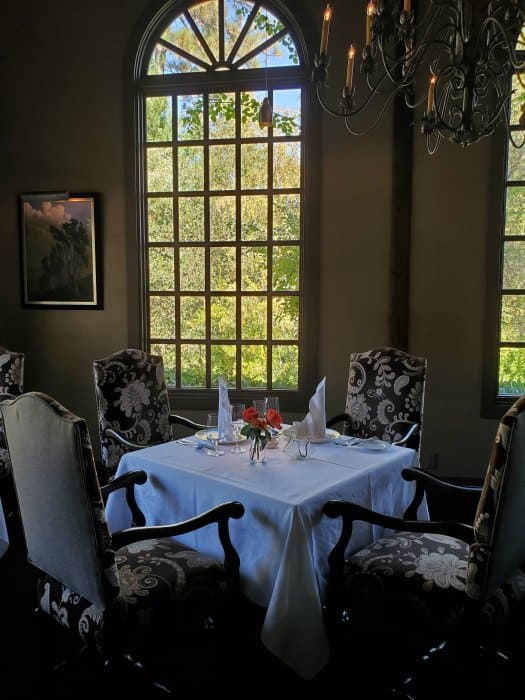 Table covered in white linen tablecloth, and place settings next to fancy chairs with a chandelier above. 