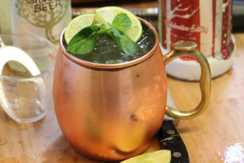 Classic Moscow Mule cocktail in a copper mug with mint and lime wedges next to a knife, lime wedge, vodka and ginger beer bottle. 