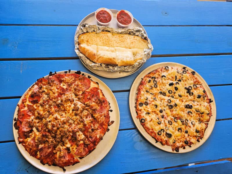 two pizzas next to garlic cheese bread with marinara sauce on a blue picnic table