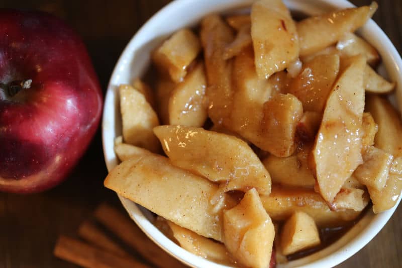 Fried apples in a white bowl next to cinnamon sticks and a red apple 