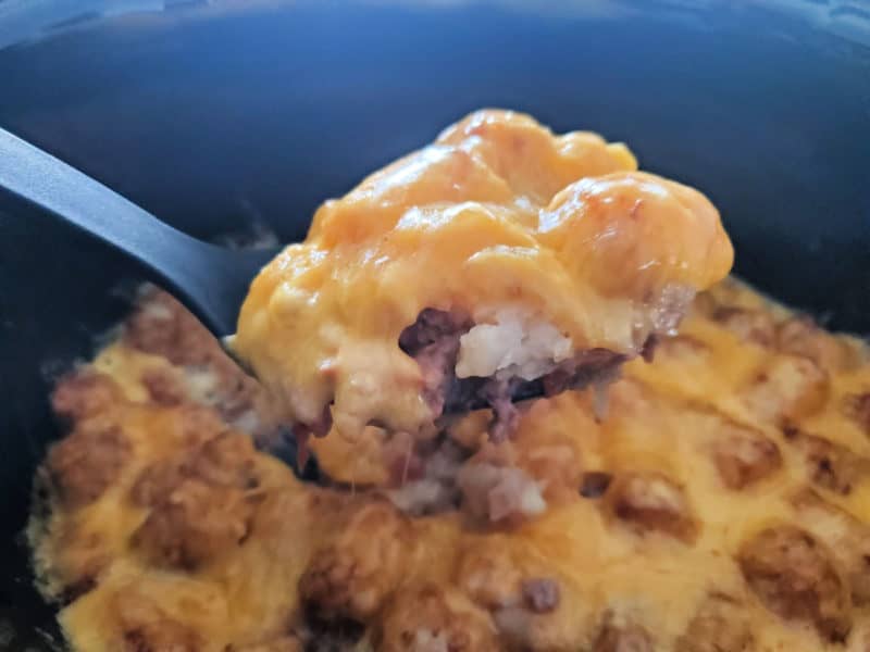 Spoon holding cheesy tater tots and ground beef over a slow cooker bowl