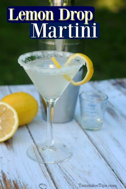 Lemon Drop Martini in a clear glass martini glass with a lemon twist and sugared rim. Cocktail shaker and a couple of lemons behind the cocktail with a white and green background. 