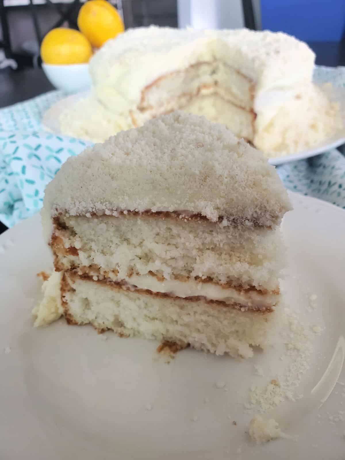 Slice of lemon cake with the rest of the cake behind it next to a bowl of lemons