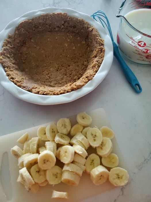 Nutter butter crust in a pie dish next to a whisk, measuring cup, and sliced bananas on a cutting board. 