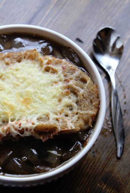 Crockpot French Onion soup in a white bowl with a silver spoon next to it on a wood background. 