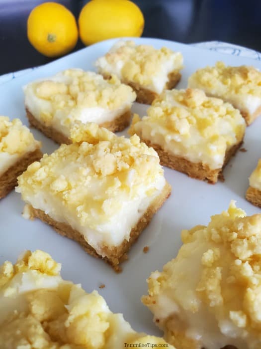 lemon cheesecake bar squares on a white platter with lemons in the background. 