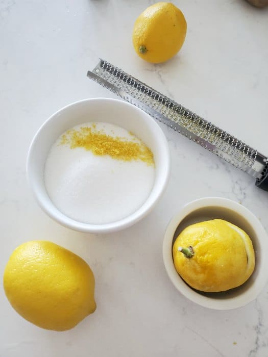 lemon zest over a bowl of sugar, zester, and lemons on a white counter. 