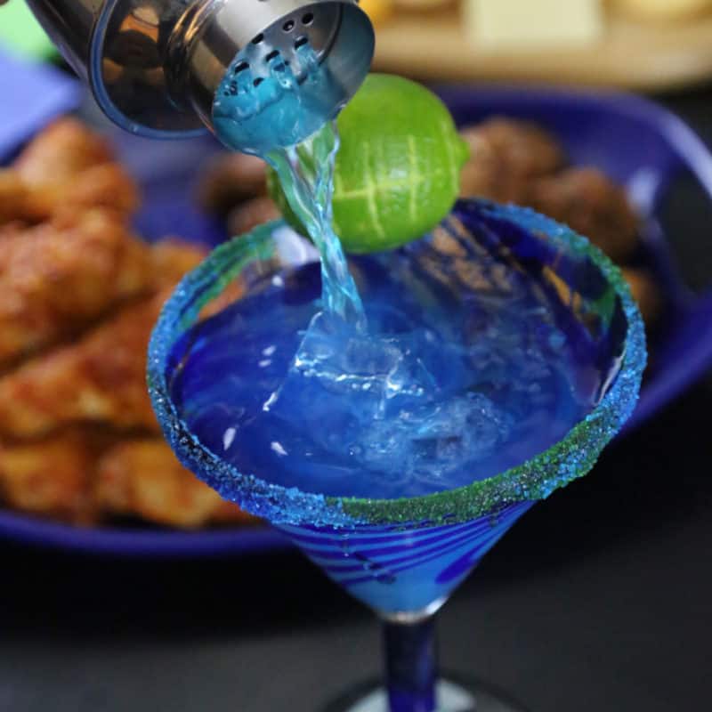 Cocktail shaker pouring blue hawk a rita into a margarita glass with a lime cut into a football