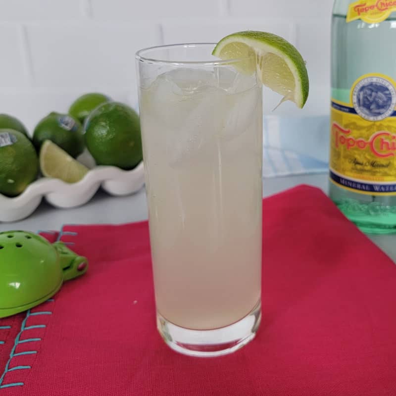Ranch Water cocktail in a tall glass next to a stack of limes, a juicer, and topo chico