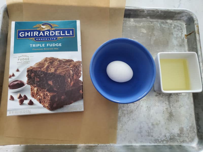 Ghirardelli triple fudge brownie box, egg in a blue bowl, and a bowl of oil on a baking sheet