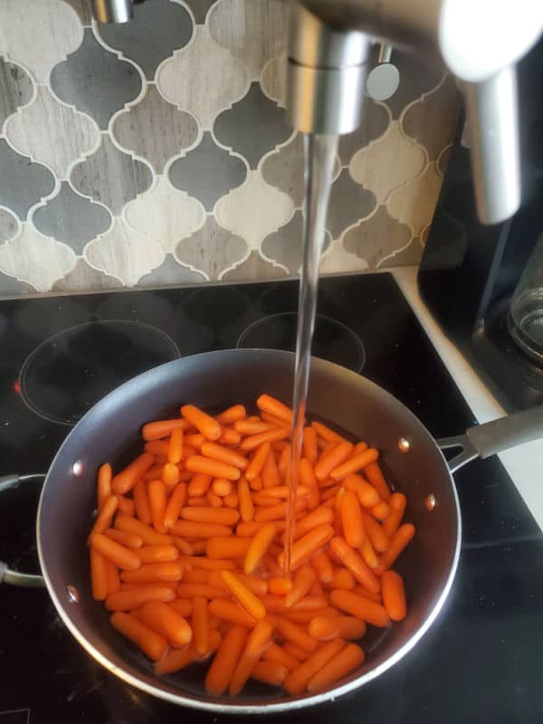 Baby carrots in a sauce pan with water flowing from a pot filler above on the stove top