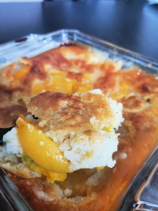 Spoon scooping peach cobbler out of a glass baking dish. 