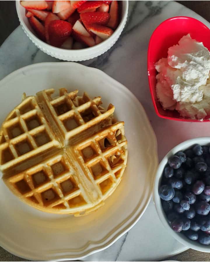 Bisquick Waffle on a white plate with strawberries, whipped cream, and a bowl of blueberries