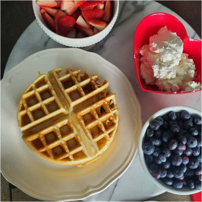 Bisquick Waffle on a white plate with strawberries, whipped cream, and a bowl of blueberries