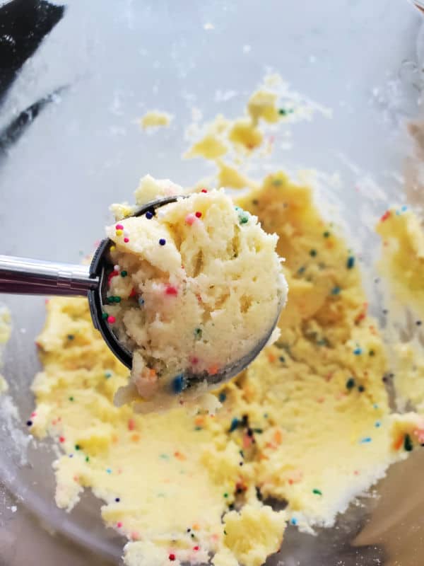 Cookie scoop with funfetti cookie dough and sprinkles held over a glass bowl