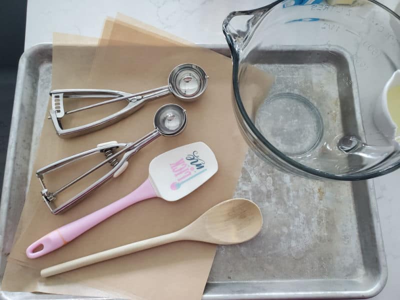 Cookie scoops, spatula, wooden spoon, parchment paper, glass mixing bowl sitting on a metal baking tray