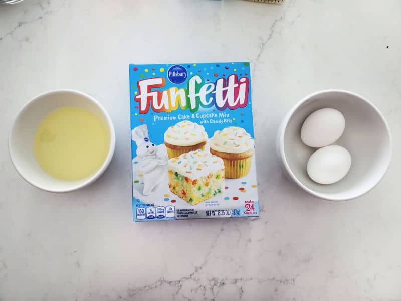 Vegetable oil, Funfetti Cake Mix box, bowl with two eggs on a white counter. 