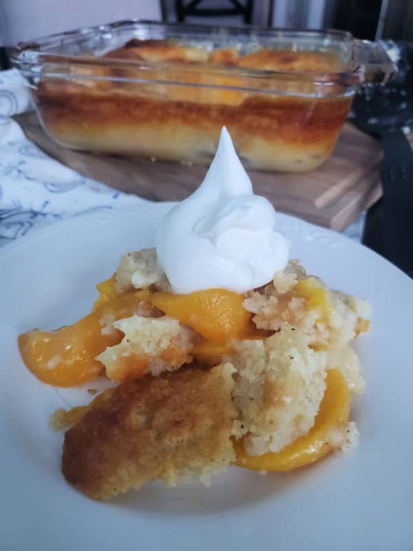 peach cobbler with a dollop of whipped cream on a white plate with a baking dish in the background