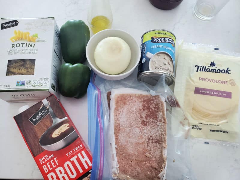 rotini pasta, green peppers, onion, olive oil, creamy mushroom soup, Tillamook provolone cheese, beef broth, and cheesesteak on a white counter. 