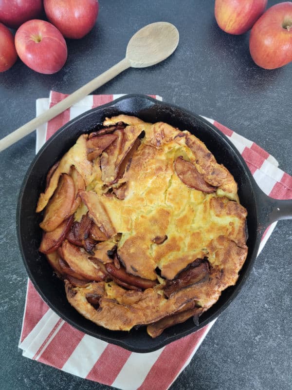 Apfelpfannkuchen German Apple Pancake in a cast iron skillet on a red striped towel next to a wooden spoon and multiple red apples