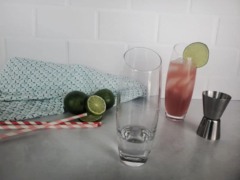 Empty highball glass with a full bay breeze drink behind it and a cocktail jigger