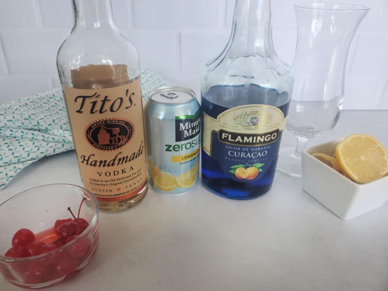 Small bowl of maraschino cherries, Tito's Vodka, Lemonade, Blue curacao, hurricane glass, and lemon wheels on a white counter. 