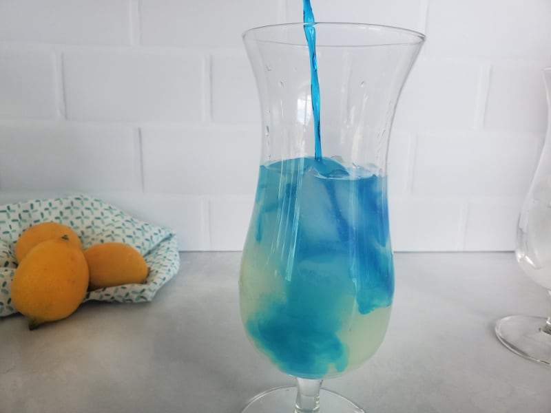 Blue liquid pouring into a hurricane glass over lemonade with lemons in the background 