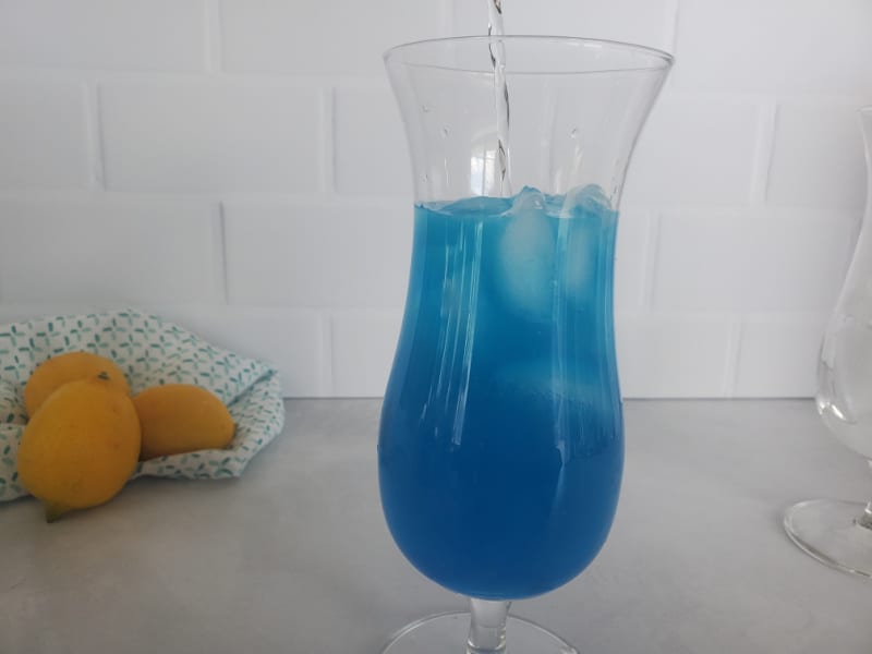 Clear liquid pouring into blue liquid in a hurricane glass next to lemons in the background. 