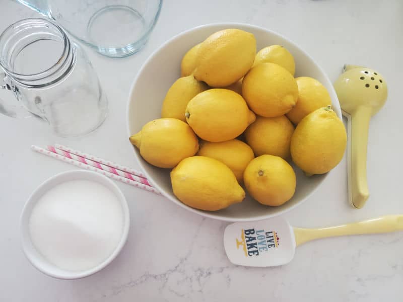 lemons in a large bowl next to a mason jar glass, bowl of sugar, spatula, and lemon squeezer