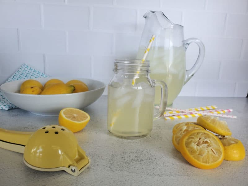 lemonade in a pitcher and a mason jar glass near a bowl of lemons, lemon juicer, and squeezed lemons