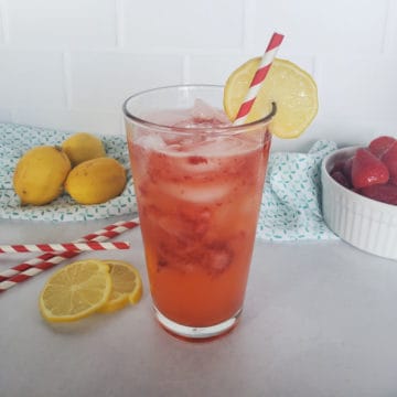 freckled lemonade in a glass with a lemon wheel garnish. lemons and striped straws in the background.