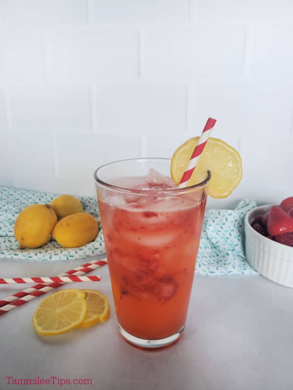 strawberry lemonade in a glass with a lemon wheel garnish. lemons and striped straws in the background.