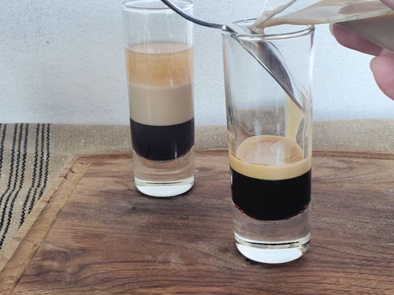 Baileys pouring over the back of a spoon into a shot glass onto dark liquid