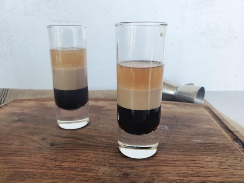 Two layered cocktail shots on a wooden board with a jigger in the background. 