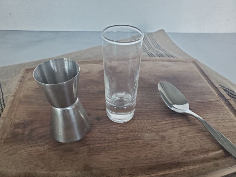 Jigger, shot glass, and spoon on a wooden cutting board. 