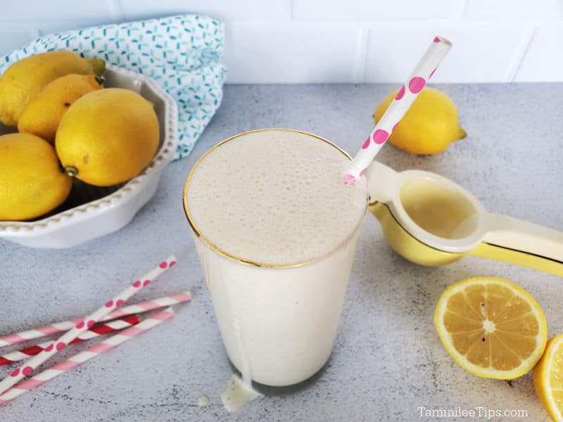 Frosted lemonade in a tall glass with a paper straw next to a bowl of lemons, paper straws, cut lemons, and a lemon squeezer. 