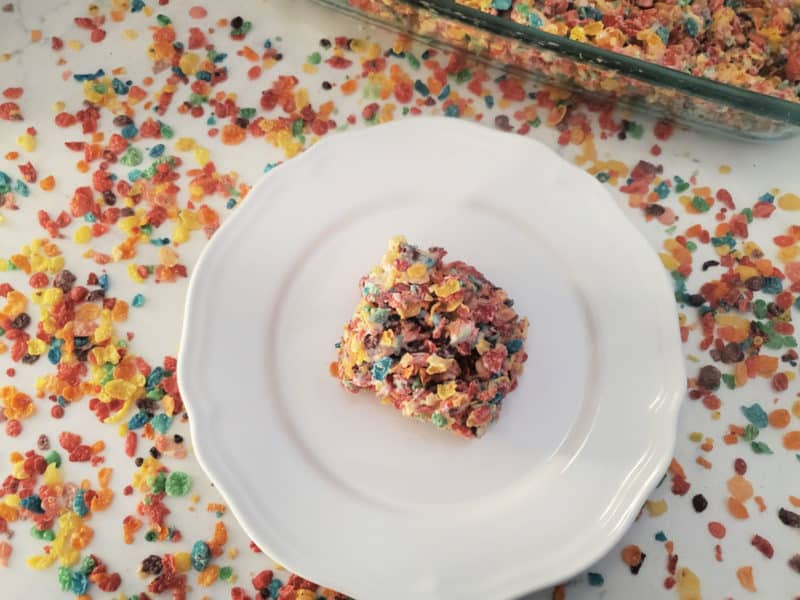 Square fruity pebbles rice crispy treat in the center of a white plate surrounded by cereal and the baking dish