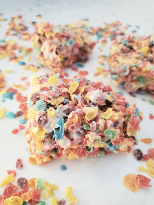 Squares of Fruity Pebbles rice crispy teats on a white counter with additional fruity pebbles cereal