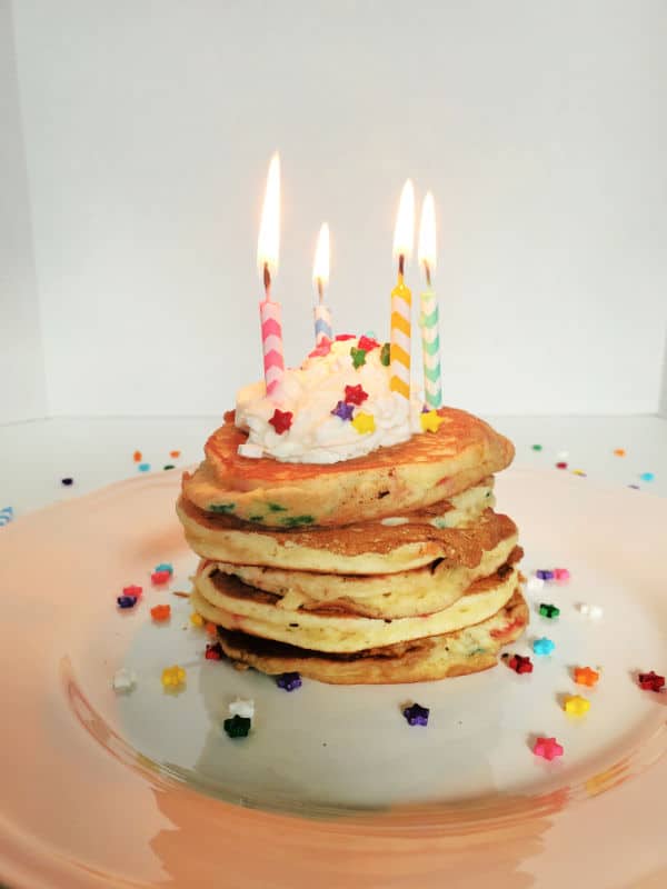 Stack of funfetti cake mix pancakes with lit birthday candles, whipped cream, and sprinkles on a white plate