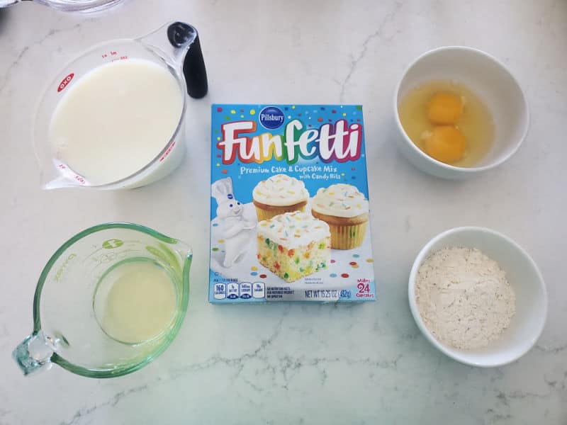 milk, oil, eggs, and Funfetti Cake Mix box on a white counter. 