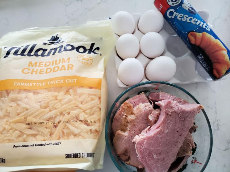 Shredded cheddar cheese, eggs, crescent roll, and ham in a bowl sitting on a white counter. 