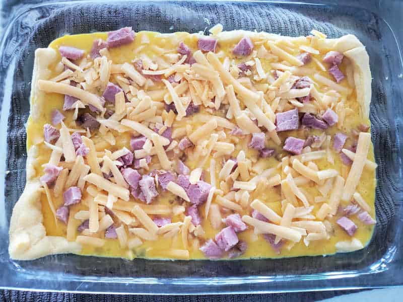 crescent roll topped with egg, ham and shredded cheese in a glass baking dish. 