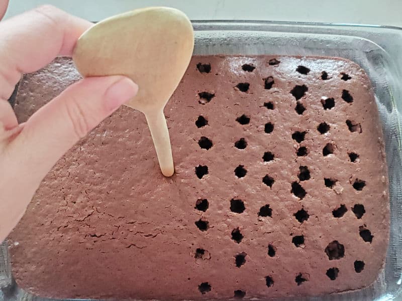 Wooden spoon poking holes in a chocolate cake in a glass baking dish. 