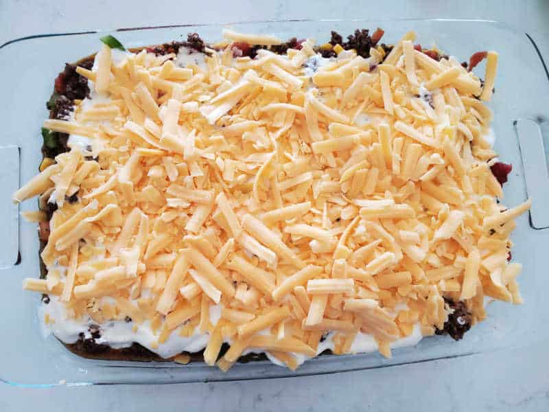 shredded cheddar cheese over ground beef, sour cream, and vegetables in a casserole dish
