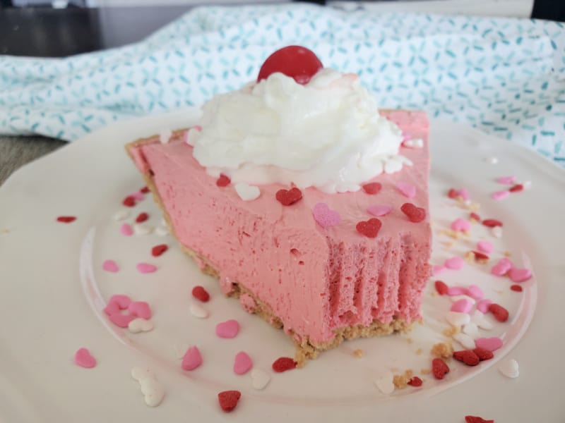 Slice of Kool-Aid Pie on a white plate with whipped cream garnish, maraschino cherry, and sprinkles